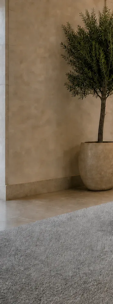 A plastered wall, a potted olive tree, a tiled floor and the edge of a grey carpet in a large entrance space